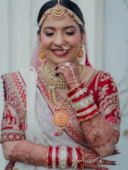 This candid shot captures the bride's shy smile and the intricate details of her look. The makeup is soft yet defined, highlighting her eyes and complementing the traditional red and white bangles (chooda).