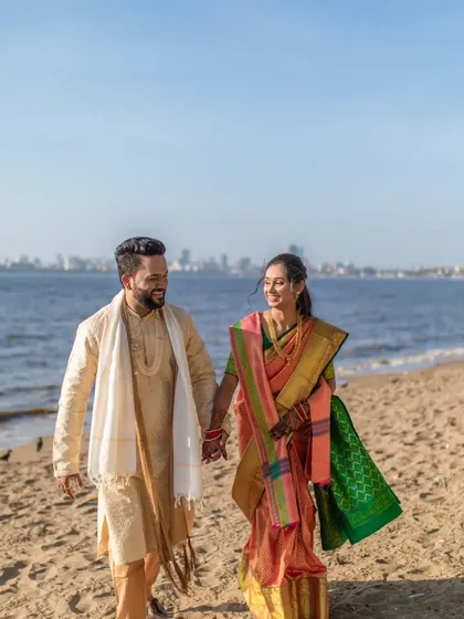 A full-length shot of the couple walking on the beach, capturing the sea, the city skyline, and their happy moment.