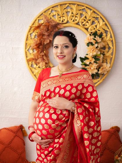 A close-up portrait capturing the radiant smile of a mother-to-be dressed in a traditional red and gold saree. The details of her hair and jewelry add to the elegance of the shot.