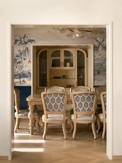 A view into a classic dining room, framed by a doorway. We used natural wood tones for the carved dining chairs and table, set against a beautiful blue-and-white toile wallpaper that wraps the space in artful serenity.