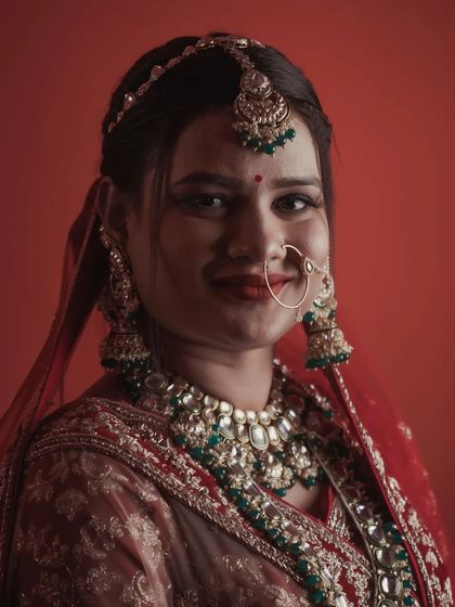 A classic bridal portrait against a simple red backdrop. The direct, gentle smile of the bride showcases her happiness, while the lighting beautifully illuminates her green and red kundan jewelry and traditional attire.