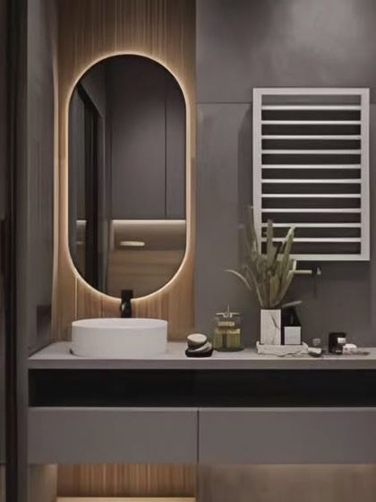 This bathroom design uses a backlit oval mirror and a sleek grey vanity to create a modern look. The heated towel rack adds a touch of functional luxury.