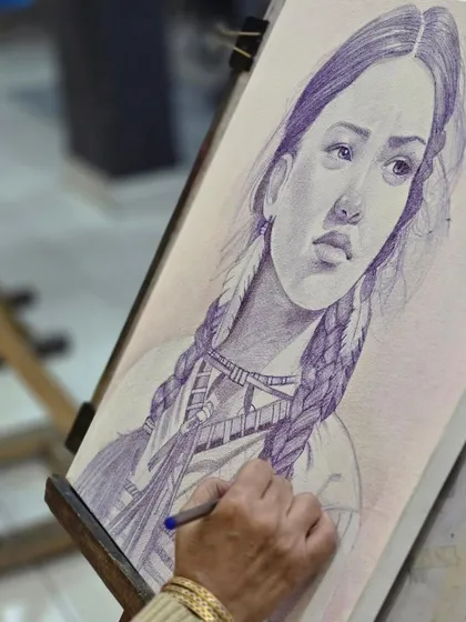 A close-up of the artist's hand and the intricate detail being added to the portrait with a ballpoint pen.