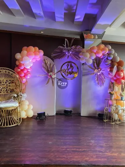 A chic and modern stage design for a baby shower. We used clean white panels, dramatic purple uplighting, and metallic palm accents to frame the "Oh Baby" sign. The iconic peacock throne and asymmetrical balloon arches complete this stylish setup.