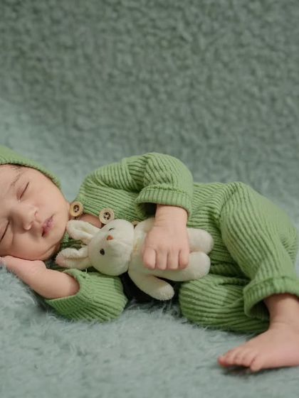 A close-up of a sleeping newborn in a cozy green ribbed outfit, cuddling a small white bunny toy on a soft green blanket.
