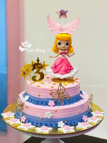 A beautiful two-tier doll cake for a 3rd birthday. The cake is decorated in pink and blue with golden butterflies and stars, topped with a winged doll figure.