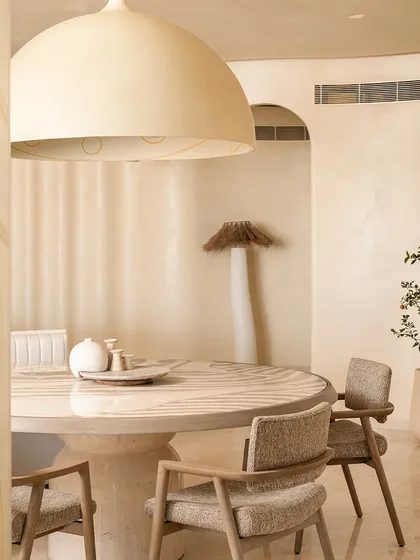 A cozy corner in the 'Soft Serve' apartment, featuring a round dining table under a large dome light. The space is designed with soft curves and a neutral palette to evoke a sense of calm and tranquility.