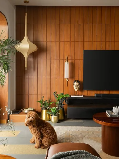 This living space is a study in modern Indian luxury. A feature wall of vertical teak veneer panels brings warmth and texture, complemented by a statement brass pendant light. The flooring, a mix of polished and unpolished Kota stone with Jaisalmer marble inlays, adds intricate detail and a connection to heritage materials.