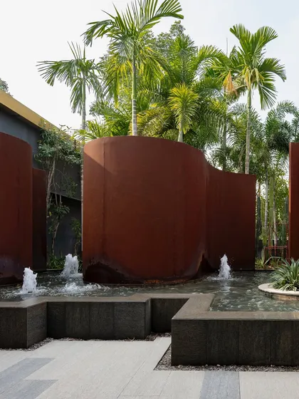 A wider view of the KEUS water feature, showing the interplay of curving Corten steel walls, bubbling fountains, and tall palm trees. The design is both sculptural and functional, creating a dynamic public space.