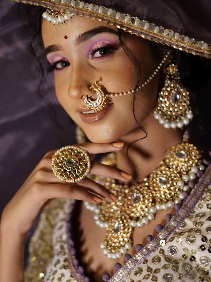 A model wearing a white and purple lehenga with a gold and pearl Kundan set. The large finger ring is part of the complete set available for rent.