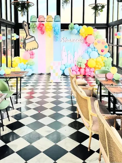 A full view of a bright and colorful art-themed party, perfect for unleashing creativity. The decor features a pastel rainbow backdrop with paint drip effects, a large paintbrush prop, and balloon garlands that look like paint spilling from buckets, all set against a stylish black and white checkered floor.