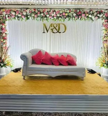 A simple and chic stage for an engagement or reception. The decor includes a white curtain backdrop, a floral border with the couple's initials (M&D), and a clean, elevated platform.