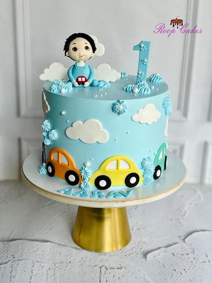 A fun car-themed cake for a first birthday. This light blue cake is decorated with colorful cars, fluffy clouds, and a handcrafted fondant figure of the birthday boy.