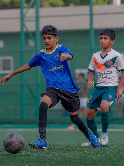 A player in a blue jersey drives forward with the ball, showing composure and control under pressure from an opponent. This is a perfect example of where talent meets tenacity.