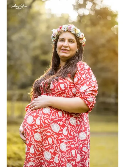 A classic maternity portrait taken outdoors. The mother-to-be, dressed in a vibrant red print dress, gently holds her baby bump, radiating warmth and happiness.