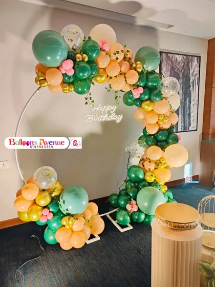 A refreshing green and peach balloon arch on a circular frame, accented with gold balloons and a 'Happy Birthday' neon sign.