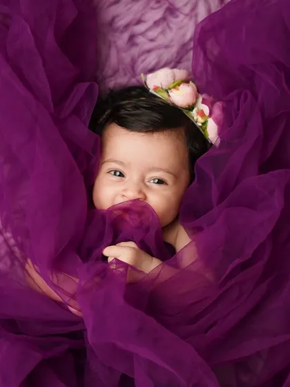A baby girl peeks out playfully from behind a swirl of purple fabric. Her mischievous smile is absolutely captivating.