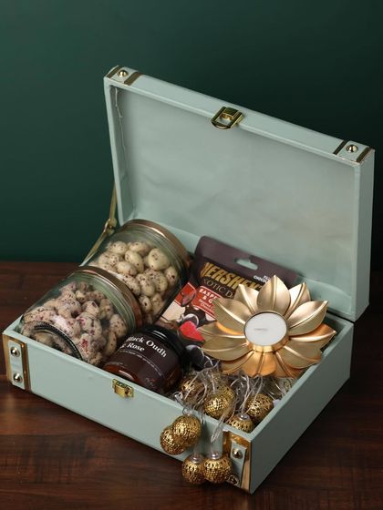 This elegant trunk-style box in a pastel green is filled with premium goodies. It includes two jars of coated nuts, a jar of black oudh and rose, a golden flower diya, and Hershey's chocolate.