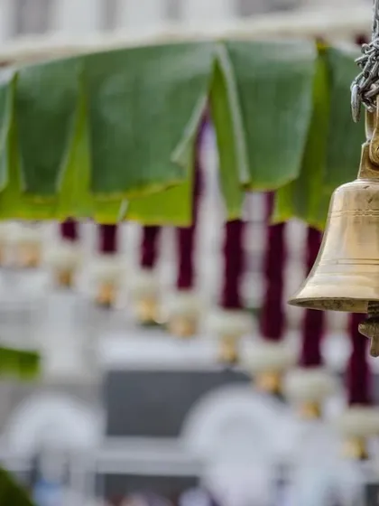 A close-up of a brass temple bell hanging from the entrance arch, set against a backdrop of banana leaves.