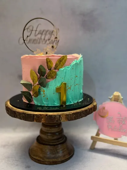 This anniversary cake features a contemporary two-tone design with pink and teal buttercream. A spray of golden leaves climbs the side, tying in with the gold '1' and the elegant 'Happy Anniversary' topper.