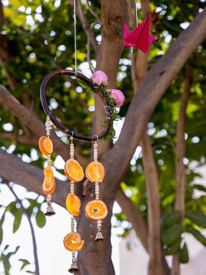 A handcrafted dreamcatcher-style ornament hanging from a tree, made with a wooden hoop, dried orange slices, beads, and small bells.
