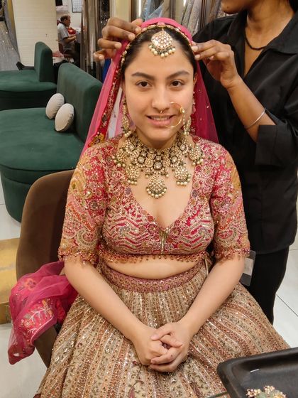 A bride getting her matha patti adjusted during a trial. She is wearing a heavy, traditional gold-toned polki set that perfectly complements her red and gold lehenga.