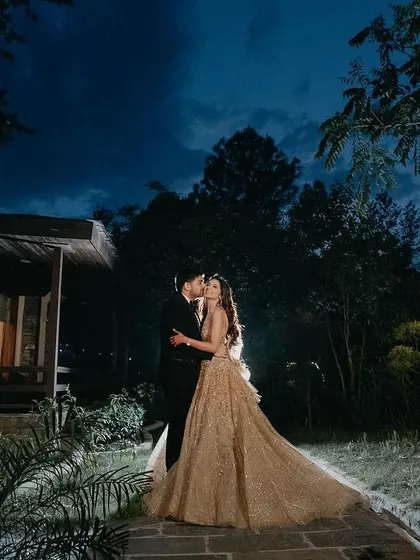 A wide shot capturing the couple in a magical, fairytale-like setting. The twilight sky and the warm lights of the cottage create an enchanting backdrop for their romantic kiss.