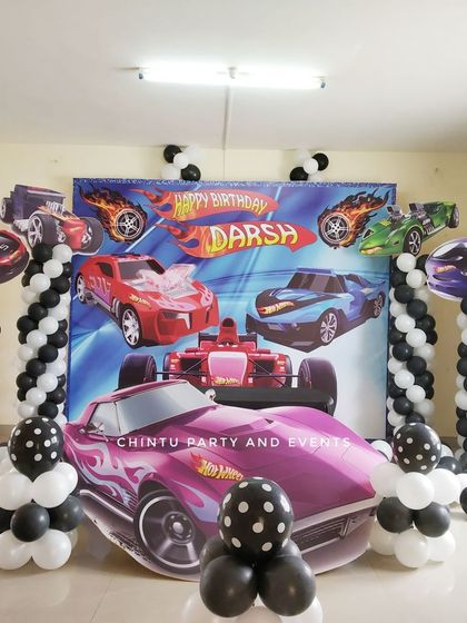 A Hot Wheels themed birthday party with a dynamic car-filled backdrop. The decor includes black and white balloon pillars and a large car cutout for photos.