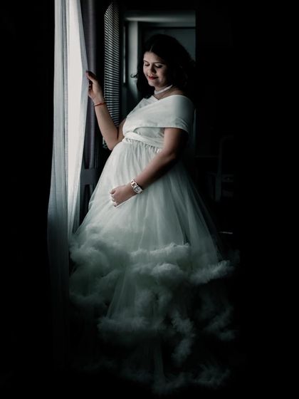 A moody and artistic window-lit portrait. The white ruffled gown catches the light beautifully, creating a high-contrast, dramatic maternity photo.