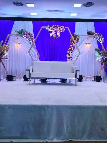 A sophisticated engagement stage with a white sofa and a backdrop of blue and white drapes. The hexagonal frames are decorated with floral arrangements.