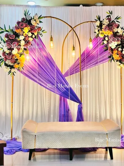 A unique engagement setup with a double-arch frame in gold. The purple drapes and colorful floral arrangements create a vibrant yet elegant look, complemented by hanging Edison bulbs.