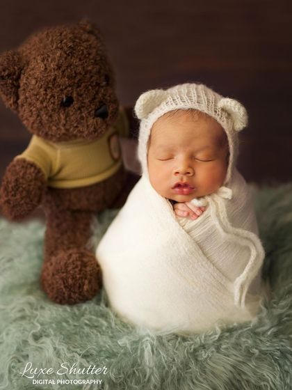 A classic newborn portrait with a baby in a white swaddle and bear-ear hat, posed next to a cuddly teddy bear.
