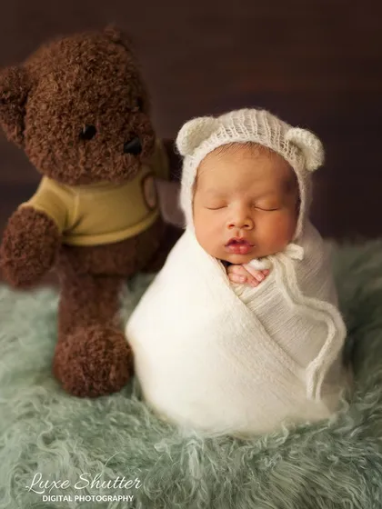 A classic newborn portrait with a baby in a white swaddle and bear-ear hat, posed next to a cuddly teddy bear.