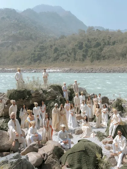 A family portrait like no other. The entire wedding party, dressed in shades of white, poses against the stunning natural backdrop of the Ganga river and mountains in Rishikesh.