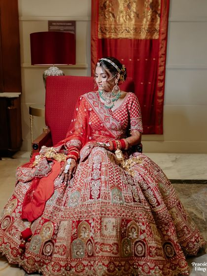 A regal portrait of the bride seated on a red chair, her elaborate lehenga arranged perfectly around her.
