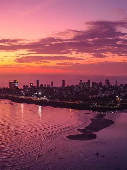 The Mumbai coastline and Marine Drive bathed in the soft, purple and pink hues of a tranquil sunset. This drone shot captures the serene meeting of the city and the Arabian Sea, with the tide creating patterns on the shore.