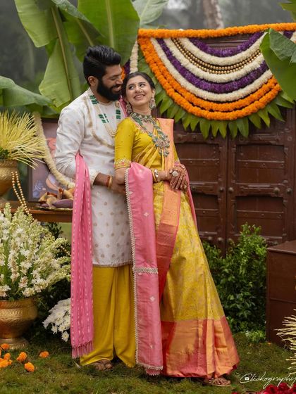 A full-length portrait of the couple, showcasing their perfectly coordinated traditional outfits. The groom's white kurta and yellow dhoti are tied together with the bride's look through a matching pink uparna (stole).