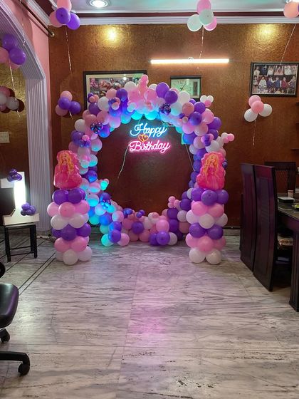 A princess-themed first birthday setup at home, featuring a circular balloon arch in shades of pink and purple, a neon sign, and a marquee number "1".