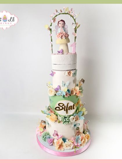 A breathtakingly tall, multi-tiered fairy cake for a magical first birthday. This elegant design features a soft green and white palette, adorned with a cascade of pastel flowers, butterflies, and a sweet fairy topper.