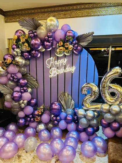 A beautiful purple and silver balloon decoration. This setup features a slatted circular backdrop, a neon sign, and a mix of matte and chrome balloons with silver leaf accents.