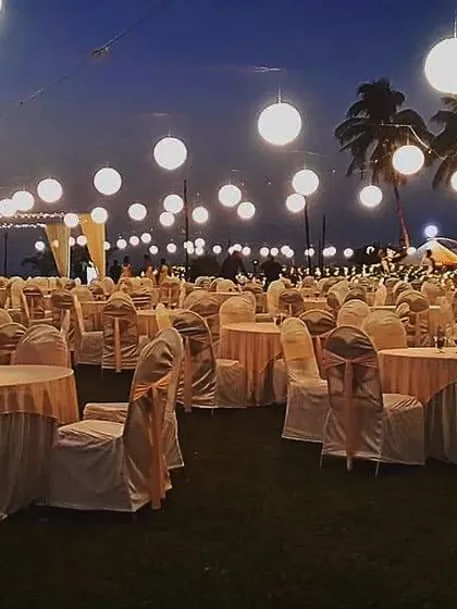 An evening view of a lawn reception setup. The large, glowing paper lanterns hanging over the guest tables create a beautiful and magical atmosphere under the night sky.