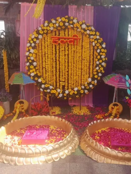 A vibrant Haldi setup with two lotus-shaped urlis filled with flower petals. The backdrop is a large ring of marigolds, decorated with small umbrellas and colorful drapes.