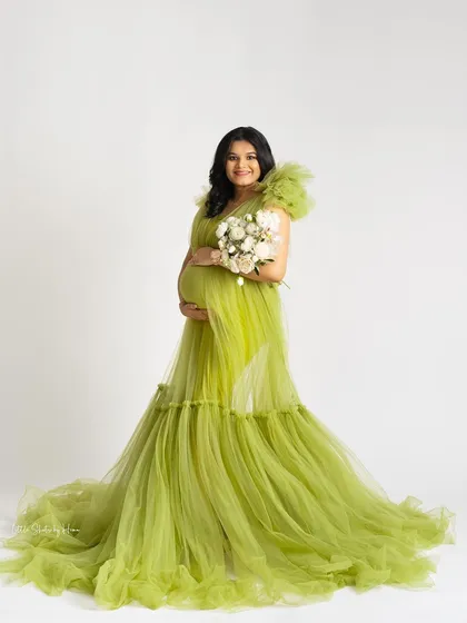 A joyful portrait of a mother-to-be in a bright lime-green gown, holding a bouquet of white flowers. She smiles warmly against a clean white studio background.