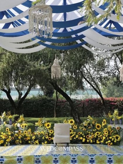 A simple yet beautiful haldi stage with a backdrop of sunflowers. The blue and white draped ceiling with macrame chandeliers adds a touch of boho-chic elegance.