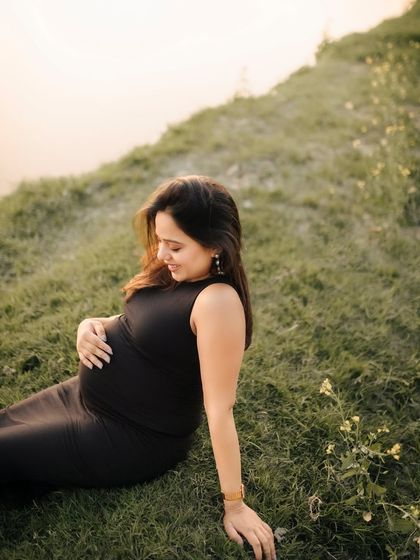 A quiet and contemplative moment as a mom-to-be sits on a grassy hill by the water. Dressed in a simple black dress, she gently holds her baby bump, lost in thought.