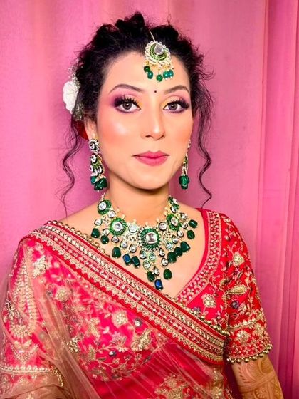A bride smiling in a red lehenga, wearing a beautiful Kundan necklace with large green stones and pearl drops. The matching maang tikka completes this classic look.