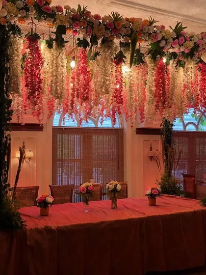 An elegant VIP dining table setup, perfect for hosting key stakeholders or award winners. The table is framed by a beautiful overhead structure with hanging floral arrangements and warm incandescent bulbs, creating an exclusive and intimate setting.