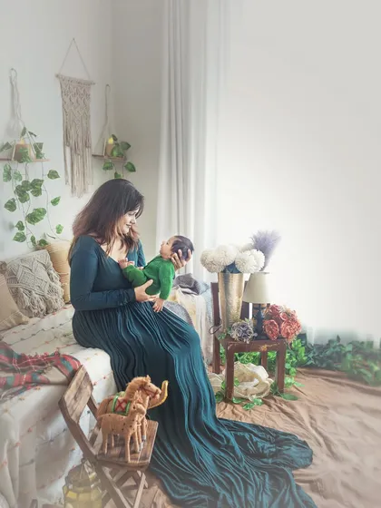 A wider shot of the new mother and her baby in the boho studio. The flowing green dress and the beautifully arranged decor create a stunning, lifestyle-focused newborn portrait.