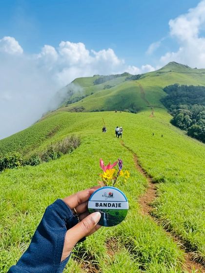 A beautiful shot of our Bandaje trek badge with wildflowers, held against the backdrop of the green trail leading up the peak.