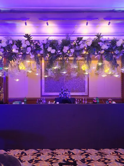A stunning bar setup featuring a ceiling installation of hanging florals and lights. The purple uplighting creates a moody and sophisticated ambiance, perfect for an evening cocktail party.